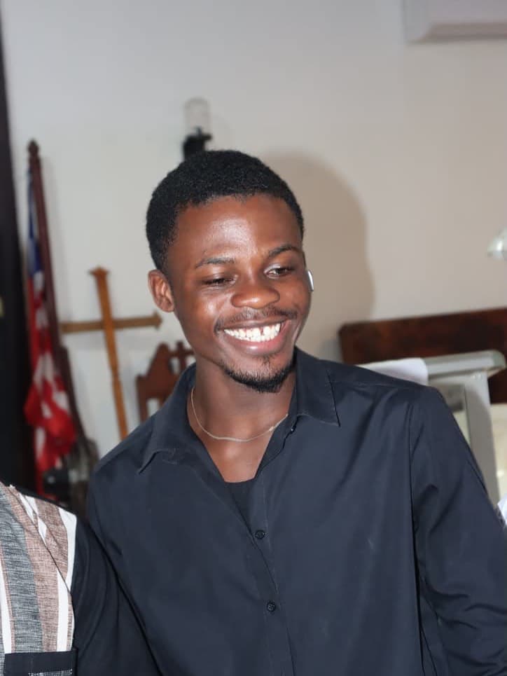 Smiling young Black man wearing a black shirt with a wooden cross in the background.