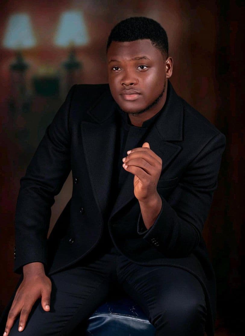 Portrait of a young Black man in a black coat, seated and looking away thoughtfully.
